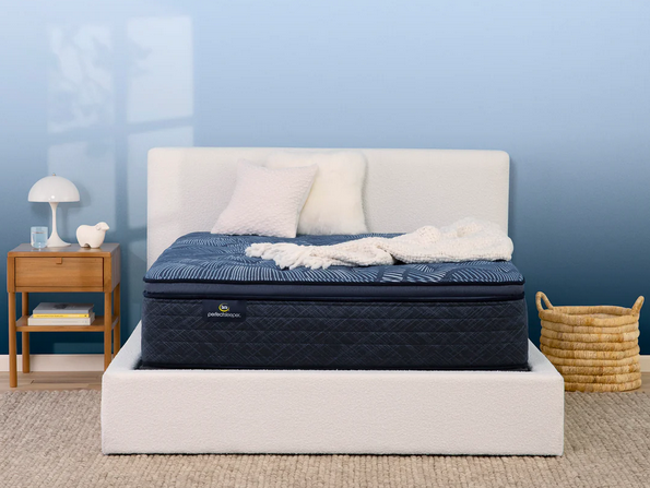 A Serta Perfect Sleeper Cobalt Calm Plush Pillow Top Mattress in a bedroom setting, showcasing the mattress with pillows on top and a nightstand beside it.